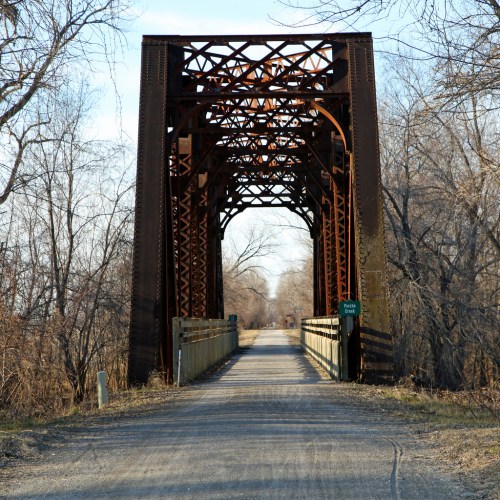 Rusty metal bridge over a gravel path, surrounded by bare trees and a clear blue sky.