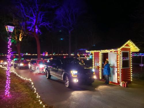 Cars lined up at a festive holiday light display, with a brightly lit booth and decorations in the background.
