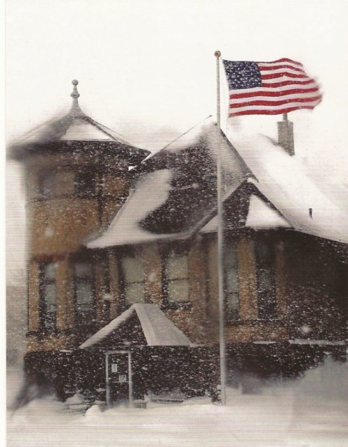 A snow-covered house with a flagpole displaying the American flag, surrounded by a winter storm.
