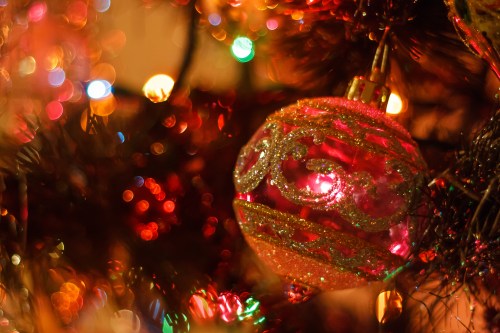 A close-up of a pink Christmas ornament hanging on a tree, surrounded by colorful, blurred lights.