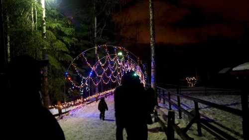 A snowy path illuminated by colorful holiday lights, with silhouettes of people walking under a light arch.