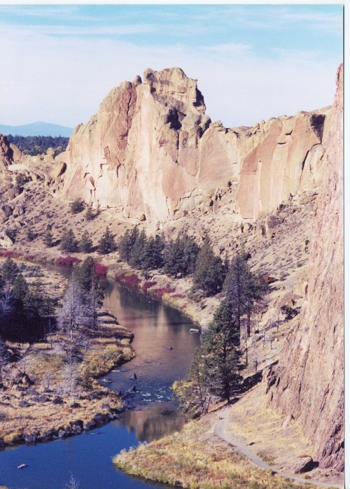 A scenic view of towering rock formations beside a winding river, surrounded by trees and open land under a clear sky.