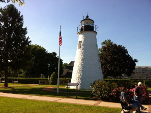 A white lighthouse with a black top stands near a flagpole and greenery, with a clear blue sky in the background.