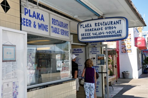 A woman walks past Plaka Restaurant, featuring a sign for homemade gyros and Greek salads, in a lively street setting.