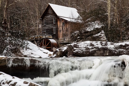 A historic wooden mill beside a frozen waterfall, surrounded by snow-covered rocks and trees in a winter landscape.
