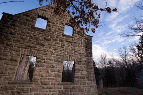 A weathered stone building with missing windows, set against a cloudy sky and bare trees in the background.