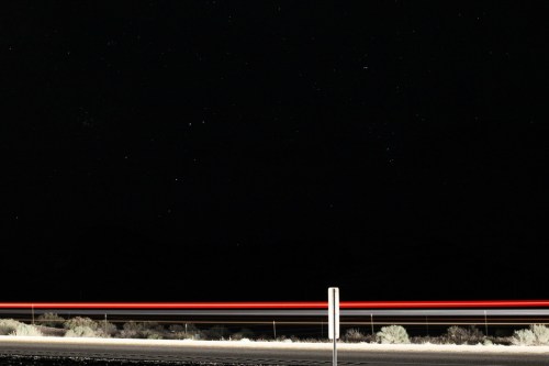 A dark night sky filled with stars, with a blurred red light streak across the bottom from a passing vehicle.