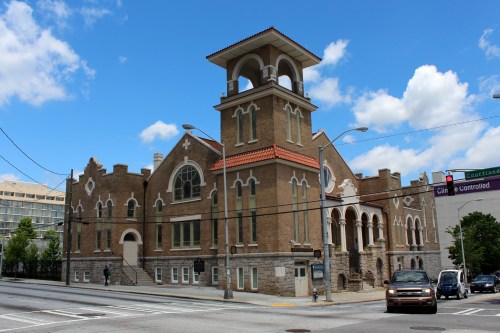 Historic brick building with a bell tower, set against a blue sky with clouds, located at a street intersection.