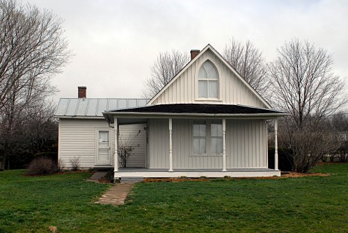 A white, Victorian-style house with a gabled roof and porch, surrounded by bare trees and a grassy yard.
