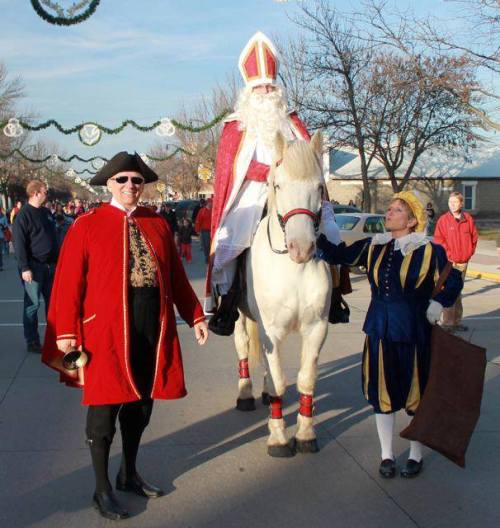 A man in a red robe and hat rides a white horse, accompanied by two people in festive costumes on a street.