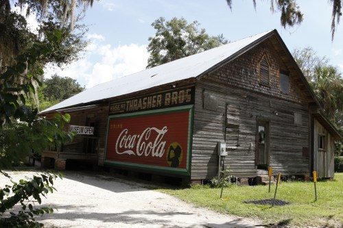 A rustic wooden building with a Coca-Cola mural, surrounded by greenery and a dirt path, labeled "Thrasher Bros."