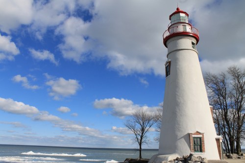 A white lighthouse with a red top stands by the shore, surrounded by trees and a calm lake under a blue sky.