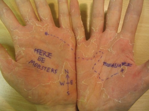 Two open hands with a drawn treasure map, featuring "HERE BE MONSTERS" and an "X" marking the treasure location.