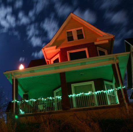 A house illuminated with green and orange lights under a cloudy night sky.