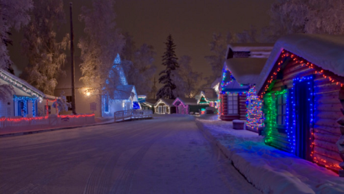 A snowy street lined with colorful, illuminated houses and trees, creating a festive winter atmosphere at night.