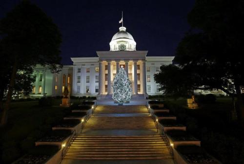 A lit Christmas tree stands in front of a grand building at night, illuminated by lights and surrounded by greenery.