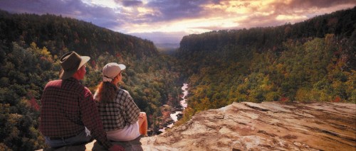 A couple sits on a rocky ledge, overlooking a lush valley and river at sunset, surrounded by colorful autumn foliage.
