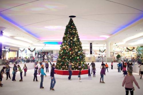 A festive ice skating rink with a large decorated Christmas tree in the center, filled with skaters enjoying the holiday season.