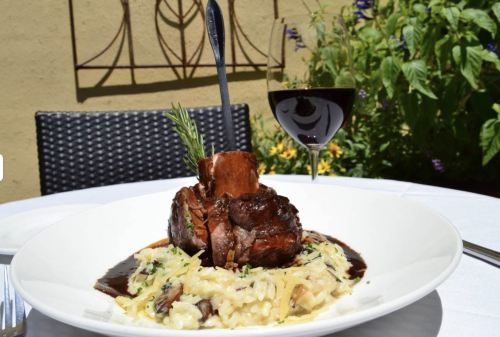 A plated dish of tender meat on a bed of creamy risotto, garnished with herbs, alongside a glass of red wine.