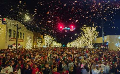 A large crowd gathers on a festive street, illuminated by holiday lights and snowflakes falling from the night sky.
