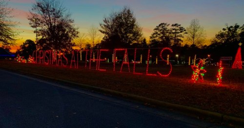 Colorful holiday lights spell out "Christmas at the Falls" against a sunset backdrop with trees in the background.