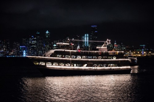 A brightly lit boat with festive decorations sails at night, with a city skyline illuminated in the background.