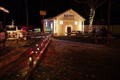 A festive scene featuring a small building labeled "Bee City City Hall," decorated with lights and surrounded by holiday displays.