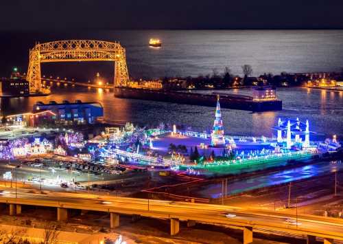 Aerial view of a festive waterfront with colorful lights, a decorated tree, and a bridge illuminated at night.