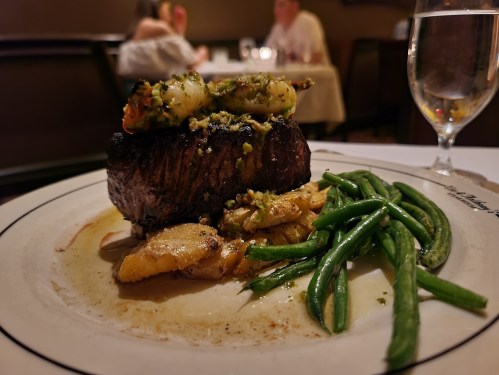 A plated meal featuring a grilled steak topped with shrimp, served with green beans and roasted potatoes.