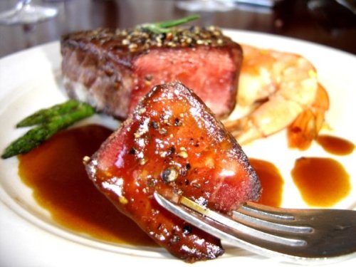 A close-up of a fork holding a piece of seared meat, accompanied by shrimp, asparagus, and a rich sauce on a plate.