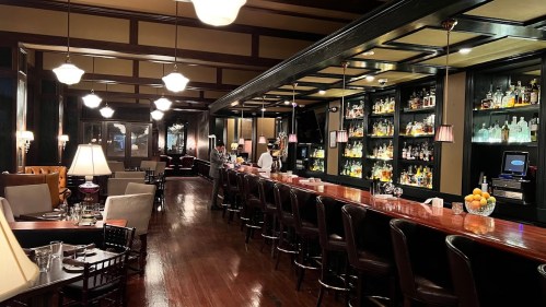 A dimly lit bar with a long wooden counter, shelves of liquor, and elegant seating. Warm ambiance with lamps and decor.