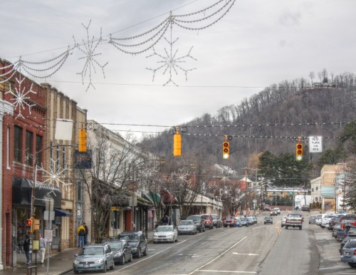 A small town street decorated with snowflake lights, featuring traffic signals and cars parked along the road.