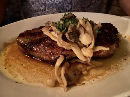 A juicy steak topped with sautéed mushrooms and herbs, served on a white plate with a savory sauce.