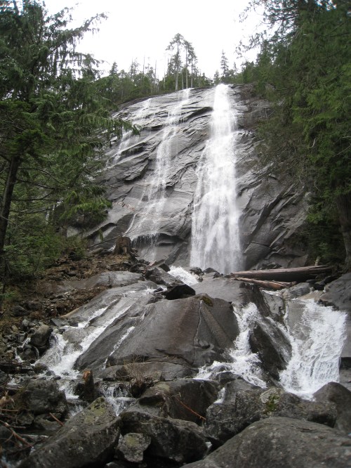 A cascading waterfall flows down a rocky cliff, surrounded by lush green trees and a rocky terrain.