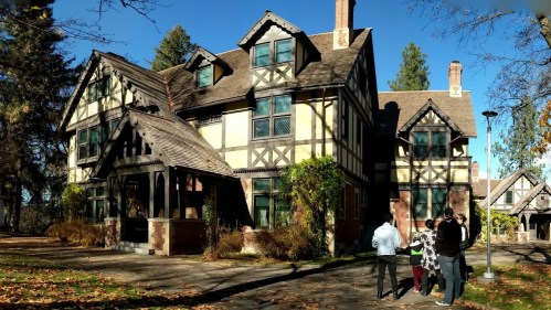 A large, historic house with a mix of architectural styles, surrounded by trees and a group of people in front.