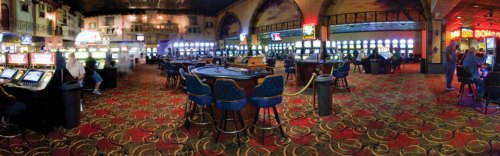 A panoramic view of a casino interior featuring slot machines and gaming tables, with patrons seated and playing.