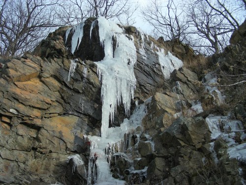 Icicles hang from a rocky cliff, surrounded by bare trees and a winter landscape.