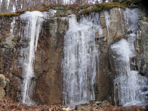 Icicles and ice formations cascade down a rocky cliff, surrounded by bare trees and earthy tones.
