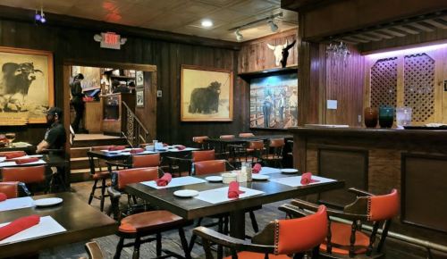 Interior of a rustic restaurant with wooden decor, empty tables set with red napkins, and a bar area in the background.