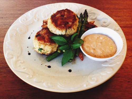 Two golden crab cakes on a plate with asparagus, grilled vegetables, and a small bowl of sauce.
