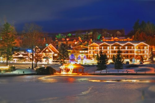 A winter scene featuring a cozy lodge illuminated with holiday lights, surrounded by snow and a frozen lake at dusk.