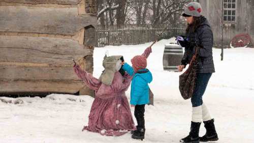 A woman and a child interact with a snow-covered figure in a winter setting, surrounded by snow and wooden structures.