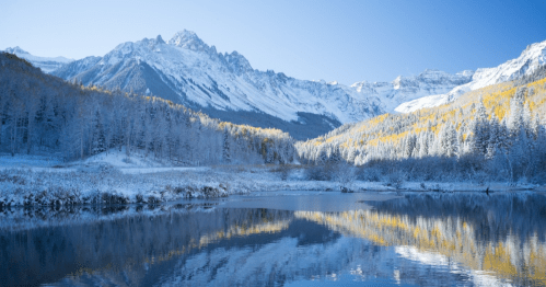 Snow-covered mountains reflect in a calm lake, surrounded by golden autumn trees under a clear blue sky.