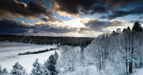 A snowy landscape with trees, a river, and dramatic clouds in the sky, illuminated by sunlight breaking through.