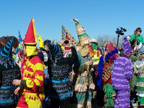 A vibrant crowd in colorful costumes and masks celebrates a festival under a clear blue sky.
