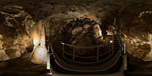 A panoramic view of a cave interior with stalactites, stalagmites, and a walkway illuminated by soft lighting.