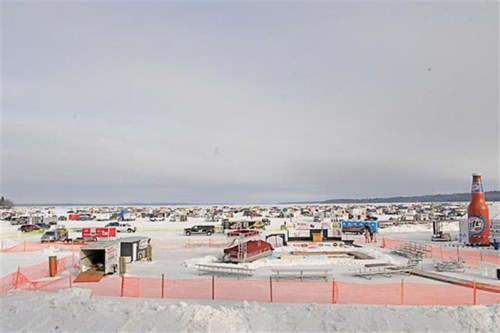 A snowy landscape filled with ice fishing huts and vehicles, with a cloudy sky overhead.