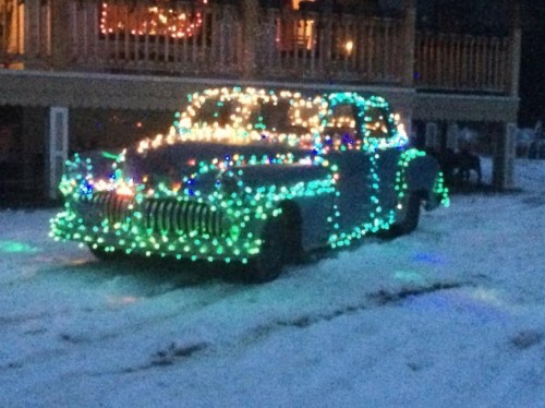 A vintage car covered in colorful Christmas lights parked on a snowy driveway.