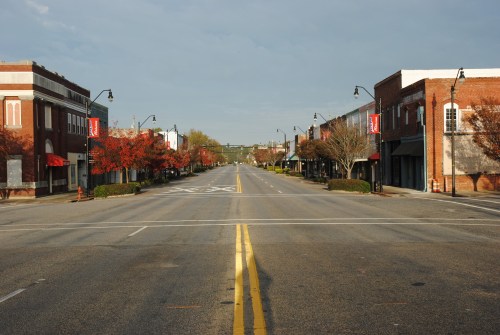 A quiet, empty street lined with trees and storefronts, showcasing autumn colors under a clear sky.