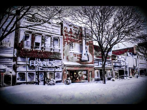 Snow-covered storefronts line a quiet street, with trees bare and a wintery atmosphere enveloping the scene.